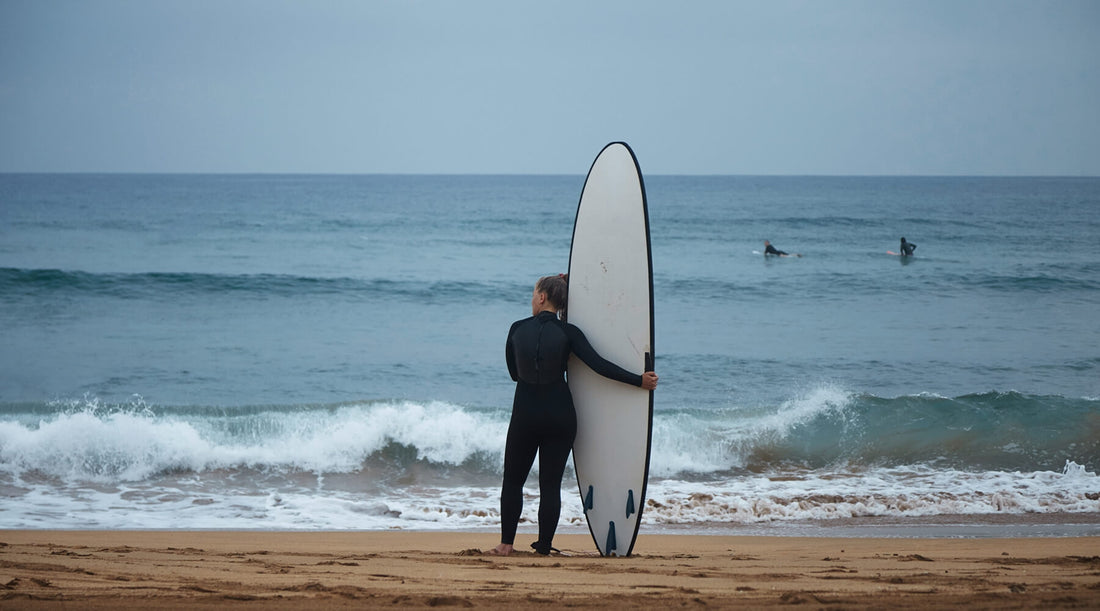 A young woman surfer, wearing a wetsuit, hugs her longboard while gazing out at the ocean and prepares for a challenging winter surfing session