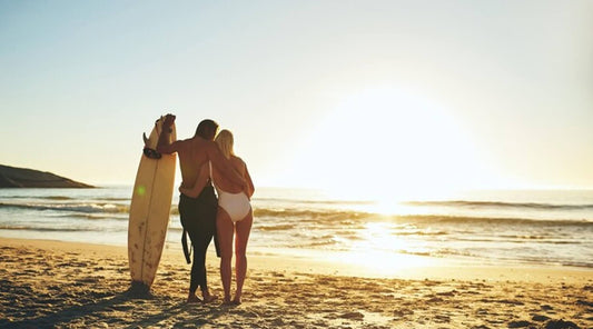 A couple holding hands on a beach at sunset, the man wears a wetsuit and hold a surfboard