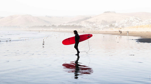 Wide-shot surfer wearing a surfing suit and holding a red surfboard, enjoying his surf trip this fall 2024