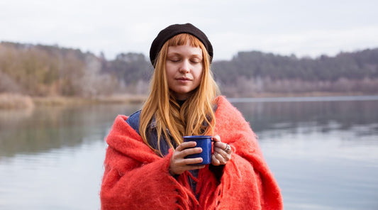 A young woman drinks coffee from a cup while sitting on the shore of a lake, preparing for a cold water swim